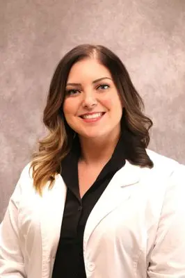 Professional woman with long brunette hair smiling at camera, wearing white blazer over black top against neutral background