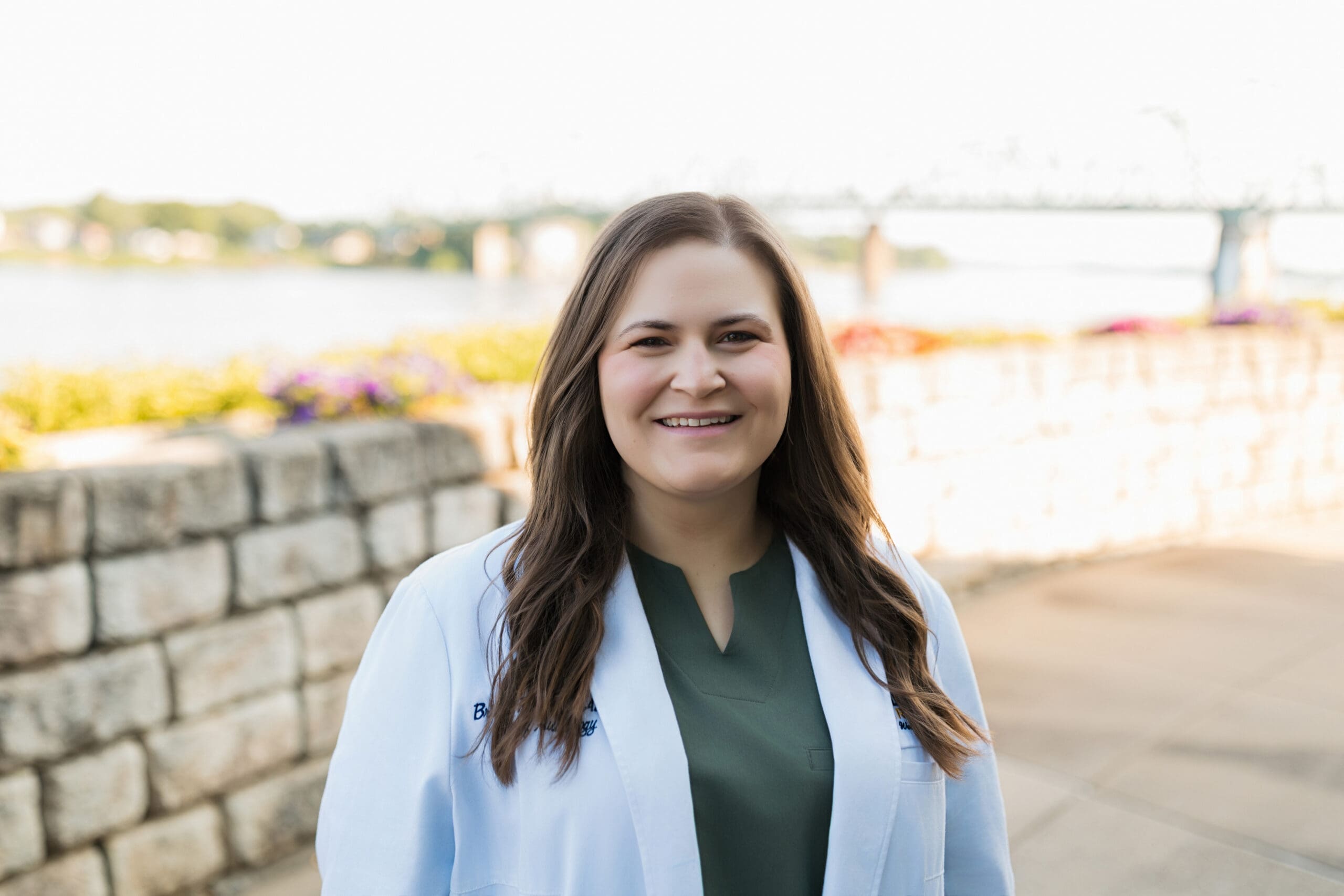 Woman in white lab coat and green scrubs smiling at camera with riverfront and flower gardens visible behind her