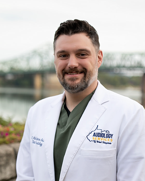 Professional man in white medical coat with audiology practice logo, smiling at camera with river and Cincinnati cityscape in background