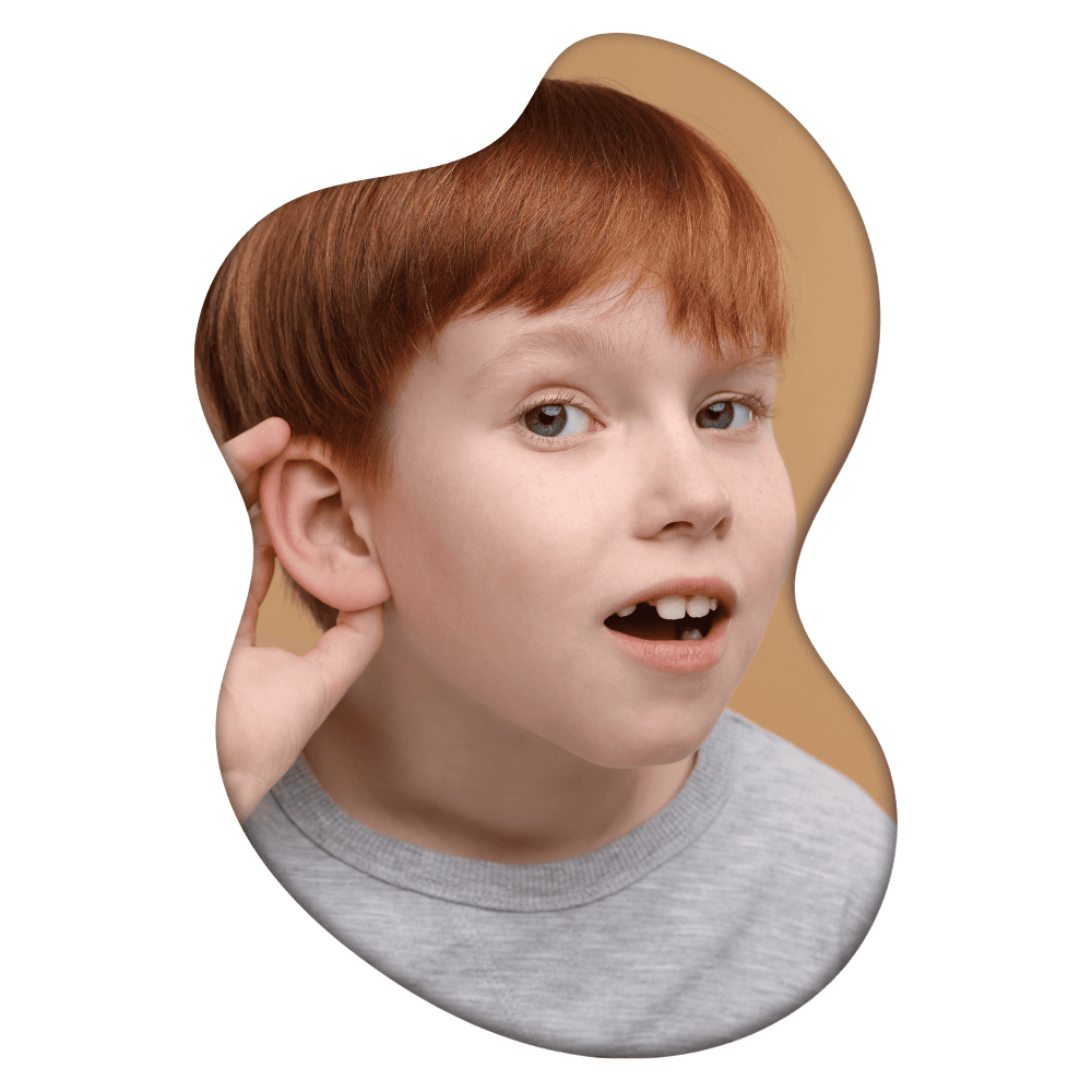 Young boy with reddish-brown hair and surprised expression, hand near ear, wearing gray shirt against tan background