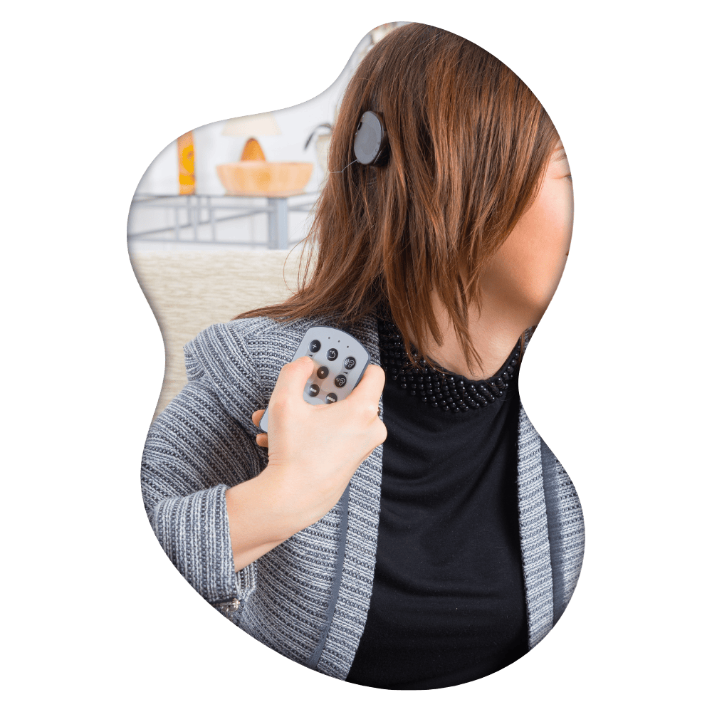Woman holding remote control facing away, wearing gray cardigan over black studded top, with blurred interior setting behind her