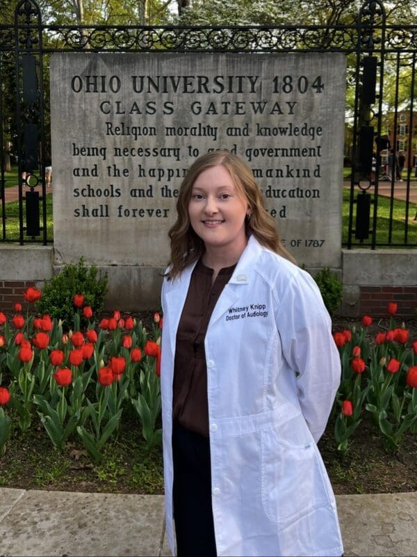 Woman in white medical coat standing in front of Ohio University 1804 Class Gateway monument with red tulips, smiling at camera