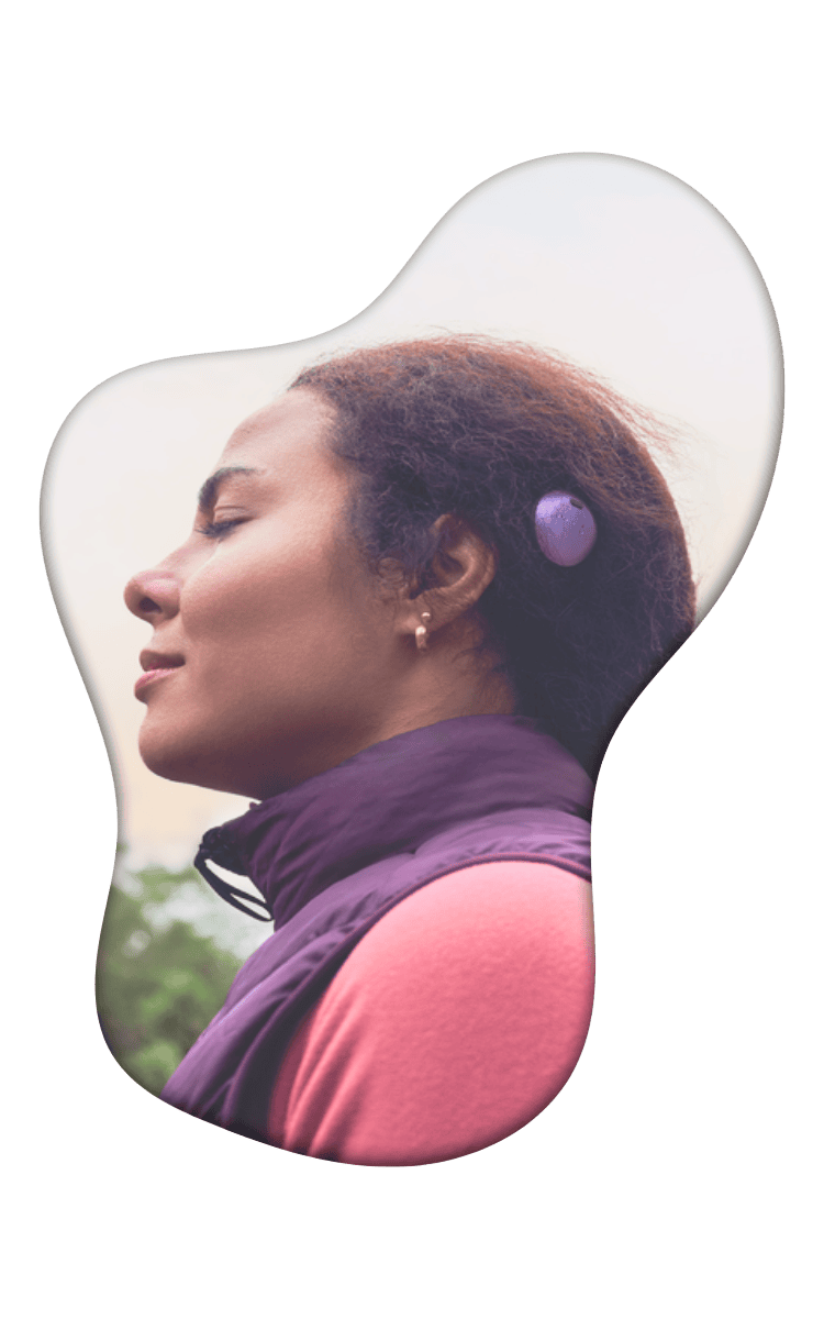Woman wearing a purple jacket and hearing aid device looking upward outdoors with blurred green background